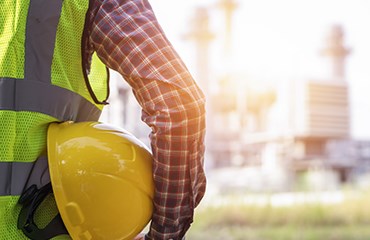 Worker looking at factory wearing hi vis jacket and holding yellow protective helmet