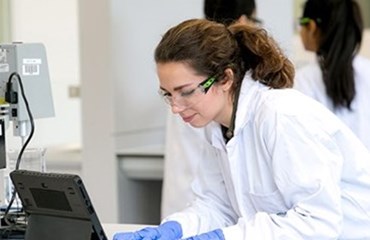Scientist wearing white lab coat protective goggles and blue plastic gloves looks at testing machine