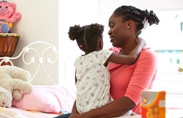 Mother cuddles child while sitting on the child's pink bed 