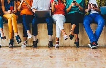 Eight people sitting on the edge of a stage looking at their phones and one in the middle looking at laptop