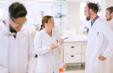 Four scientists wearing protective goggles and white lab-coats working in a Scholl lab